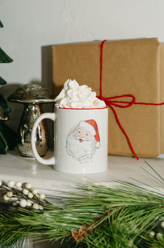 A Santa Coffee Mug filled with a whipped cream-topped drink sits on a mantel, with a brown gift box tied in red string, silver mushroom ornaments, and festive greenery arranged behind it.