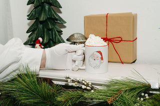 A gloved hand holds the Santa Coffee Mug filled with whipped cream. The mug sits on a white surface next to pine branches, a red-string-wrapped gift, and a green tree decoration.