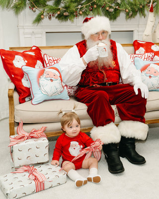 A baby in a red outfit sits among gifts as someone dressed as Santa drinks from a mug on a festive couch, which features holiday decor including the Childrens Santa Pillow in Pink and garland above.