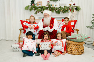 Santa Claus sits on a couch with five smiling children, one holding the Childrens Green Candy Cane Pillow, alongside a wrapped gift and festive holiday decor brightening the background.