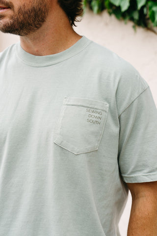 A man wears the SDS Nashville '12 South' Tee, a light gray Comfort Colors unisex shirt with a chest pocket featuring SEWING DOWN SOUTH print. The cropped photo shows his upper torso and part of his face, with greenery behind him.