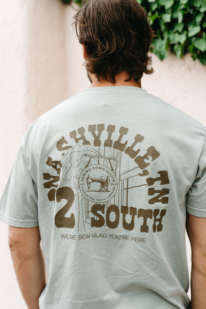 A man wears an SDS Nashville 12 South T-Shirt, light green with Were Sew Glad Youre Here and a sewing machine graphic on the back, capturing Tennessees charm amidst lush green foliage for that perfect 12 South Storefront vibe.