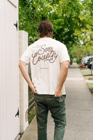 Wearing a cream-colored ‘Life is Better in Sew Country’ Tee, a brown-haired person walks on a sidewalk. “Life is better in Sew Country, Nashville, TN” is printed on the shirt, with green trees and a fence in the background.