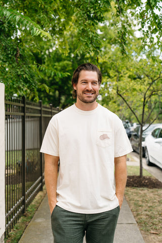 A man with brown hair and a beard stands outdoors on a sidewalk, hands in his pockets, wearing a 'Life is Better in Sew Country' Tee and green pants; trees, a fence, and parked cars appear in the background.