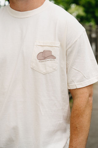 Wearing an off-white 'Life is Better in Sew Country' Tee, this unisex shirt features a chest pocket with a cowboy hat illustration and "Sewing Down South" below. The background displays blurred greenery and a fence.