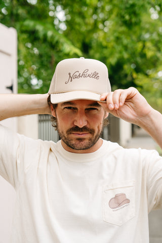 A man adjusts his SDS Nashville Rope Trucker Hat, complete with an adjustable snapback. He sports a cream T-shirt showcasing a cowboy hat graphic on the pocket. The softly blurred trees and building wall in the background accentuate his relaxed style.