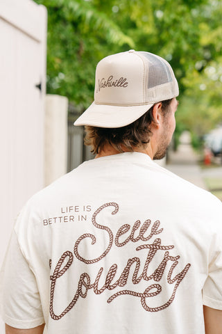 A man in a tan mesh trucker hat with "Nashville" embroidery and a white "Life is Better in Sew Country" Tee stands outdoors with green trees behind him.