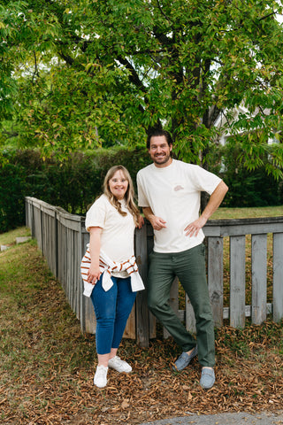 A smiling man and woman stand outdoors before a wooden fence and leafy tree. She wears jeans and a 'Life is Better in Sew Country' Tee; he pairs green pants with a light unisex tee. Both look relaxed on an autumn day.
