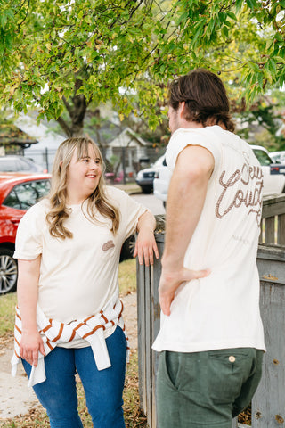 Two people chat by a wooden fence. The woman, in a cream unisex tee and blue jeans, smiles as the man leans on the fence wearing the cream 'Life is Better in Sew Country' Tee and green pants.