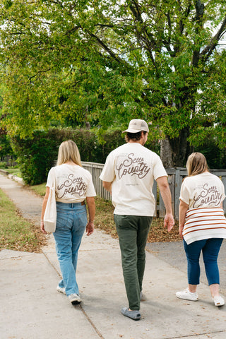 Three people walk down a sidewalk near a wooden fence and trees. Two wear matching 'Life is Better in Sew Country' Tees, while the third wears a striped shirt. Their backs highlight these popular unisex tees from Sewing Down South.