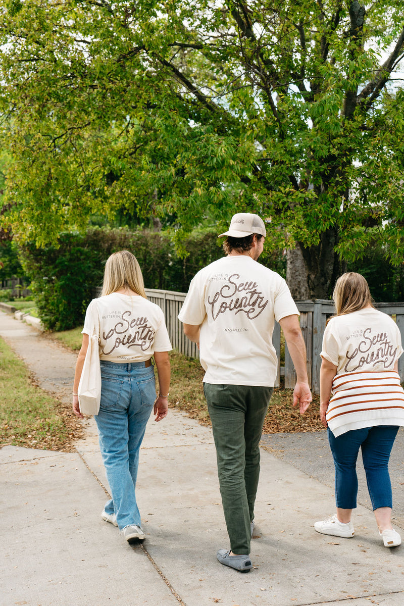 Three people walk on a sidewalk by a wooden fence, wearing cream-colored unisex tees featuring Life is Better in Sew Country on the back. The sunny day in a green, leafy area captures the essence of Americana spirit.