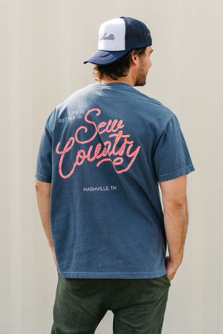 A man stands against a light wall, wearing a blue 'Life is Better in Sew Country' Tee featuring red and white "Life is better in Sew Country, Nashville, TN" text on the back. Inspired by Sewing Down South style.