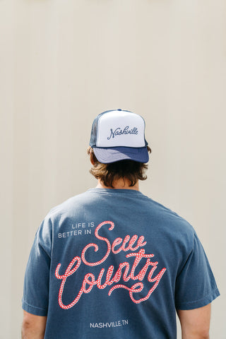 Facing away, the person wears a blue t-shirt with Life is Better in Sew Country and Nashville, TN in red script. They sport a white and blue SDS Nashville Rope Trucker Hat featuring an embroidered SDS collegiate logo and adjustable snapback for a perfect fit.