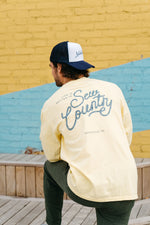 A man in a backward baseball cap and a yellow Life is Better in Sew Country Long Sleeve Tee relaxes on a wooden bench. A yellow and blue brick wall, echoing Nashvilles charm, provides the vibrant backdrop with Life is better in Sew Country and Nashville, TN printed on the shirt.