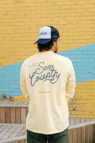 A person wearing a cream Life is Better in Sew Country Long Sleeve Tee with Nashville, TN stands confidently against a yellow brick wall with a blue stripe. Paired with a Nashville store cap, they capture the lively essence of Sewing Down South.