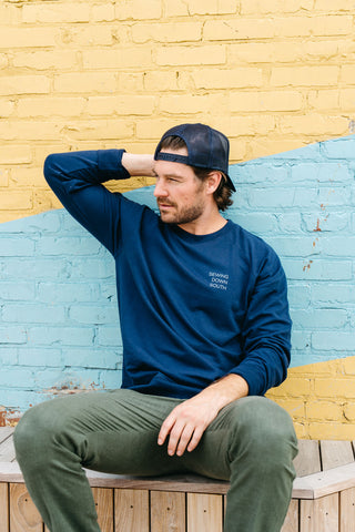 Wearing a SDS Nashville 12 South Crewneck Sweatshirt and a navy cap, a man sits thoughtfully on a wooden bench against the lively yellow and blue bricks of a Charleston storefront, adding charm to his contemplative pose.