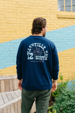 A person with brown hair, wearing an SDS Nashville 12 South crewneck sweatshirt and green pants, stands facing a colorful wall. The vibrant blue of their cozy top contrasts the yellow and blue background, reminiscent of a Charleston storefront, with wooden steps and greenery around.