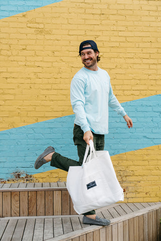 A smiling man wears the SDS Nashville Rope Long Sleeve Tee with green pants and gray shoes, carrying a large white bag as he steps playfully on wooden steps outside a Nashville store with a vibrant yellow and blue brick wall.