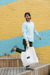 Wearing the SDS Nashville Rope Long Sleeve Shirt in blue chambray, dark pants, and a cap, a person smiles with a large white tote. They stride confidently past a yellow and blue striped brick wall on wooden planks below.