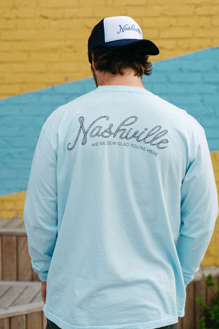 A person in a blue SDS Nashville Rope Long Sleeve Tee and a black-and-white Nashville cap stands with their back to the camera against a vibrant yellow and blue wall.