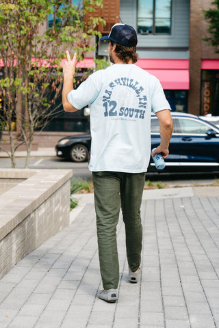 A man in a light blue SDS Nashville '12 South' Tee, green pants, and a trucker hat walks on the sidewalk holding a matching bottle and flashing a peace sign, with a car and buildings in the background.