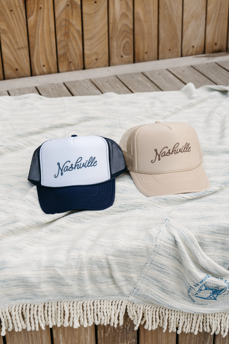 Two SDS Nashville Rope Trucker Hats, one white with an adjustable snapback and black mesh back and the other beige, featuring the embroidered SDS collegiate logo, are showcased on a patterned blanket outdoors on wooden decking.