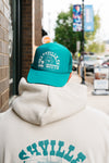 A person wearing an SDS Nashville 12 South Trucker Hat in turquoise and a white hoodie strolls down the street, framed by blurred signs and buildings.