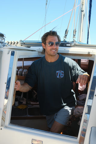 A man wearing sunglasses, The 1776 Tee unisex crewneck, and shorts stands in the doorway of a sailboat cabin, smiling and looking out on a sunny day.