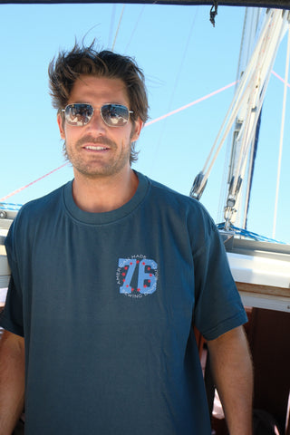 A man with tousled hair and sunglasses, wearing The 1776 Tee, smiles on a boat under bright sunlight, with ropes and sails in the background.