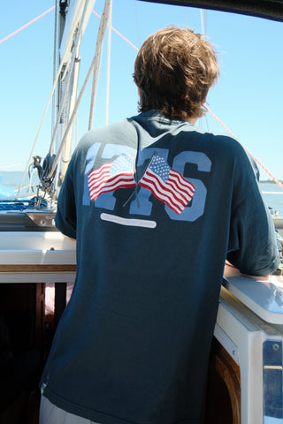 A person with brown hair, wearing The 1776 Tee emblazoned with two American flags and the numbers 1776, stands on a sailboat gazing over the water on a sunny day.