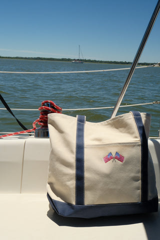 An American Flag Embroidered Tote, made of natural cotton canvas with blue handles and two flag patches, sits on a white boat seat. Behind it, water, a red rope, and a sailboat shine beneath the clear blue sky.