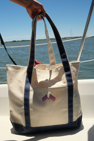 A hand holds the American Flag Embroidered Tote, a beige cotton canvas bag with navy blue handles and trim and two crossed American flags on the front, while on a boat with water and blue sky in the background.