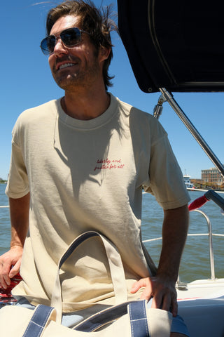 A man in sunglasses and The Loyal Liberty Tee sits smiling on a boat, holding a cream and navy tote bag. Water and buildings are visible in the background.
