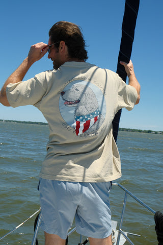 A man wearing sunglasses, The Loyal Liberty Tee with a dog graphic, and light blue shorts stands on a boat holding a sail rope, gazing at the water under a clear blue sky.