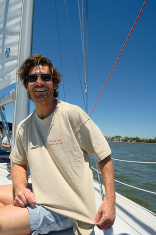 A man wearing sunglasses and The Loyal Liberty Tee is smiling while sitting on a sailboat under a clear blue sky, with water and distant buildings in the background—showcasing classic Sewing Down South style.