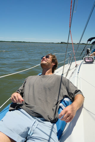 A man in sunglasses relaxes and smiles in a One Nation Tee while reclining on a sailboat under a clear blue sky, with water and a distant shoreline in the background.