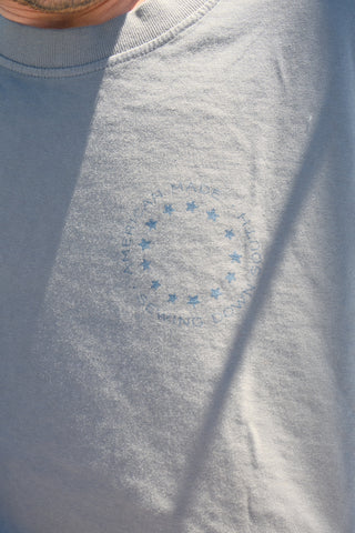 A close-up of a light blue unisex One Nation Tee by Sewing Down South, showing a faded circular star design and faint text on the chest. The lower face and neck of a person are visible in the top left.