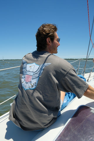 A man in sunglasses sits on a sailboat, facing away, wearing the unisex One Nation Tee with a bold eagle emblem. Water and blue sky fill the background.