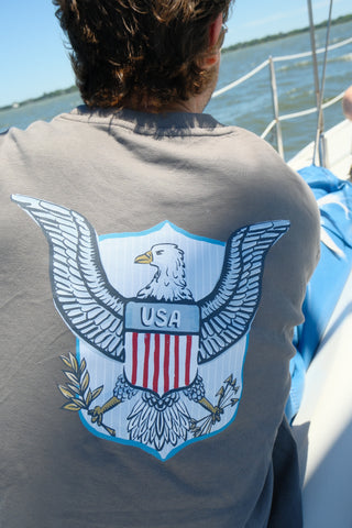 A person sits on a sailboat wearing the One Nation Tee by Sewing Down South, a unisex crewneck featuring a large USA emblem with an eagle holding an olive branch and arrows. Calm water surrounds the boat with land in the background.