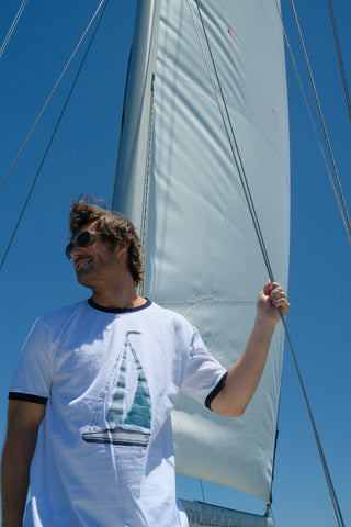 A man in sunglasses wears the Set Sail Ringer Tee, featuring a sailboat design, as he stands on a sailboat holding a rope with the sail and blue sky behind him—a perfect Sewing Down South look.