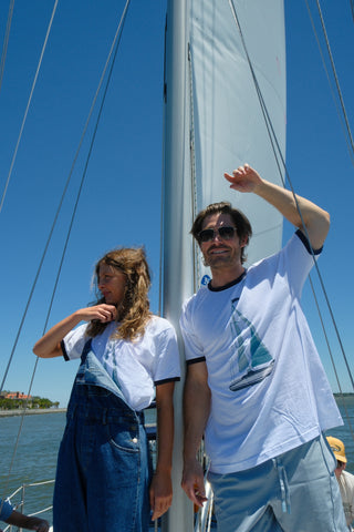 Two people in matching Set Sail Ringer Tees stand on a sailboat beneath a clear blue sky. The woman adjusts her hair while the man smiles and waves, water and shoreline behind them—a perfect Sailing Down South scene.