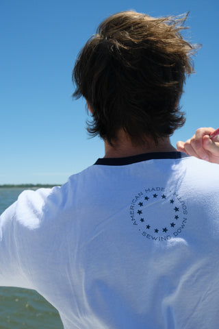 A person with brown hair faces away, wearing the white Set Sail Ringer Tee from Sewing Down South, which features a circular logo and stars on the back. The clear blue sky and water are visible in the background.