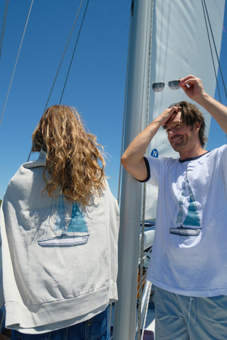 Two people stand on a sailboat under a clear sky. One faces away in a jacket with a sailboat graphic, while the other, wearing the Set Sail Crewneck Sweatshirt, smiles and lifts their sunglasses.