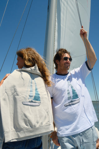 Two people stand on a sailboat under a blue sky. The woman wears the Set Sail Crewneck Sweatshirt draped over her back, while the man wears a WWWMS Tee. Both look relaxed and happy in these stylish, unisex crewnecks.