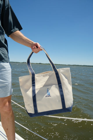 Wearing light blue shorts and a dark shirt, a person stands on a boat with water and blue sky behind, holding the Set Sail Embroidered Tote—an oversized bag featuring navy trim and a sailboat design.