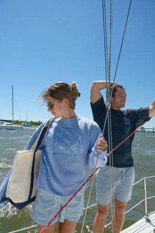 A woman in sunglasses and a light blue USA Ribbon Crewneck Sweatshirt stands on a sailboat holding a tote bag, smiling with a man in shorts and a navy shirt nearby under a clear blue sky.