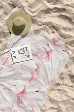 A sunhat, a magazine titled “REALTY,” and the Flamingo Beach Towel with pink flamingo prints are laid out on the sandy beach. The cotton towel has some sand on it.