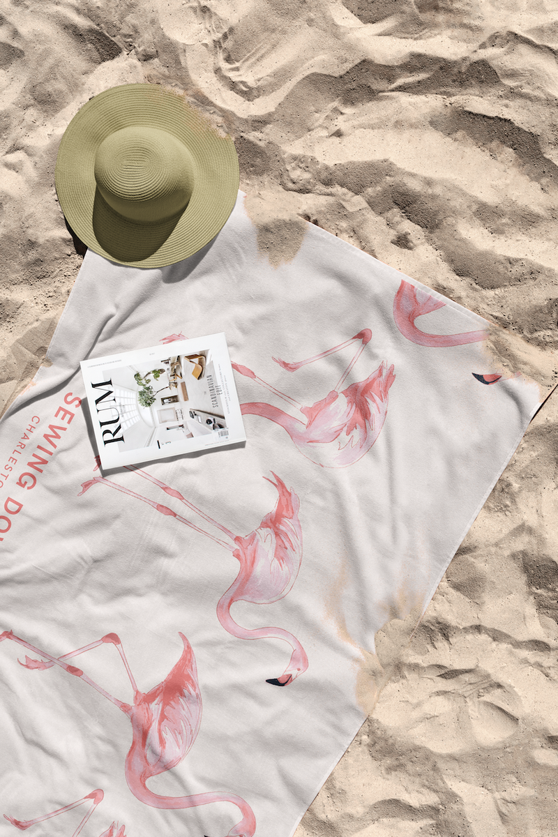 A sunhat, a magazine titled “REALTY,” and the Flamingo Beach Towel with pink flamingo prints are laid out on the sandy beach. The cotton towel has some sand on it.