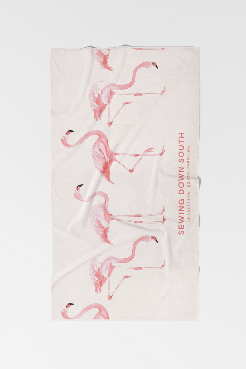 The Flamingo Beach Towel is a light-colored cotton towel featuring pink flamingos and SEWING DOWN SOUTH CHARLESTON, SOUTH CAROLINA printed vertically on the right side—a unique addition to any collection.