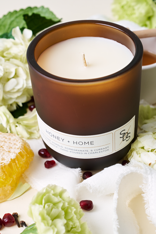 The Honey + Home Signature Candle, hand-poured in Charleston, is surrounded by green flowers, honeycomb, pomegranate seeds, and a white cloth for a cozy, fresh vibe.